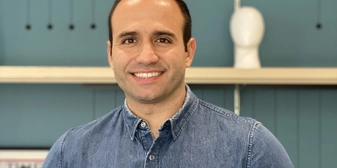 Imagem de um homem branco com uma camisa jeans. Ao fundo, uma estante azul.