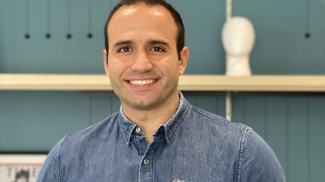 Imagem de um homem branco com uma camisa jeans. Ao fundo, uma estante azul.